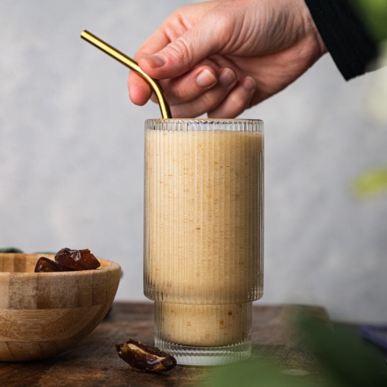 A hand holding a gold metal straw in a textured glass filled with a light brown banana smoothie, next to a wooden bowl with dates on a wooden surface.