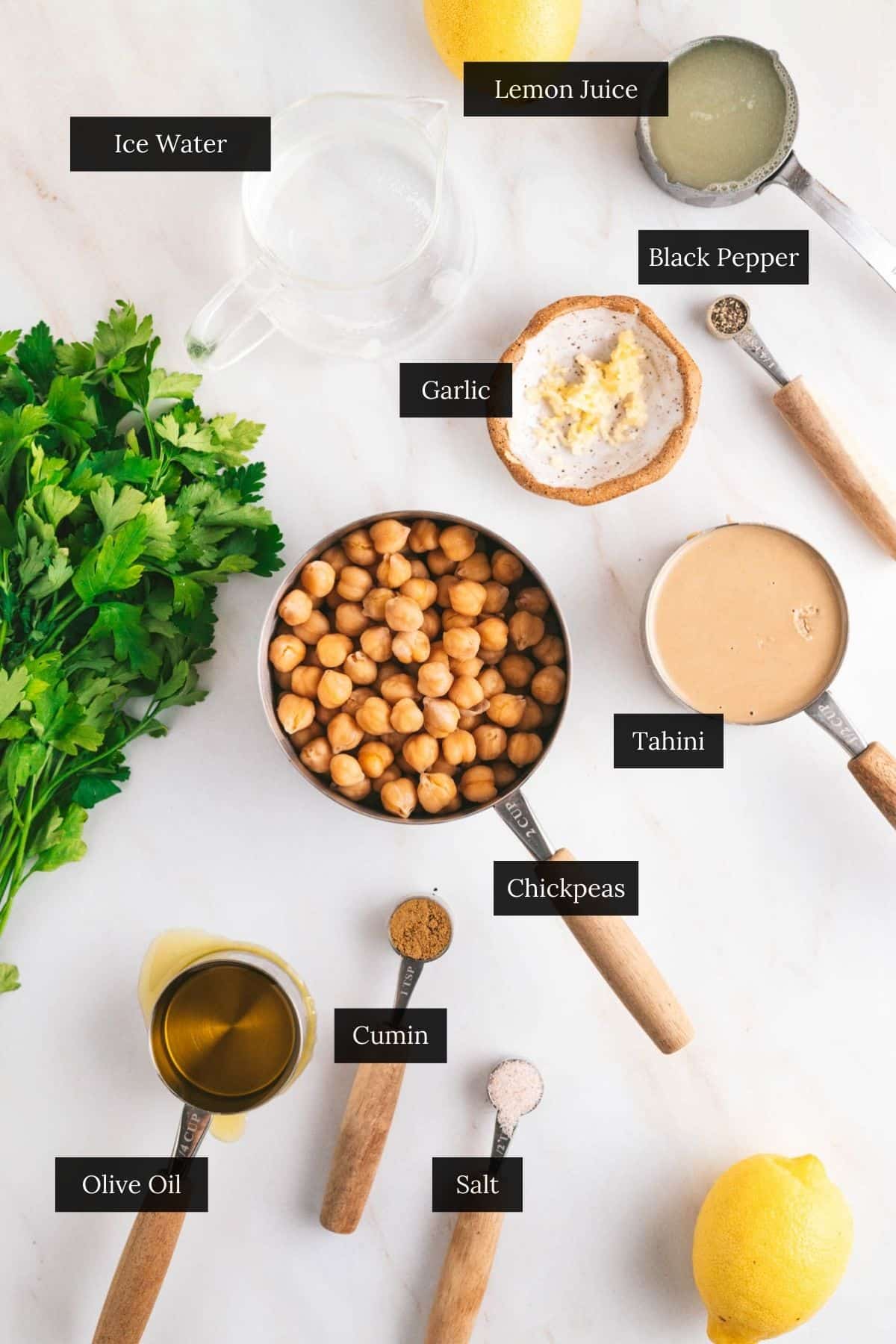For the best hummus recipe, gather ingredients like chickpeas, tahini, lemon juice, olive oil, garlic, parsley, cumin, black pepper, salt, and ice water on a white surface.