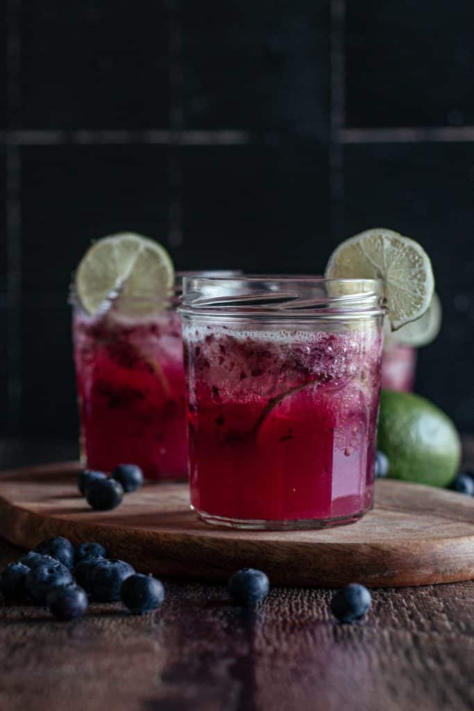 Two glasses of blueberry limeade on a wooden cutting board.