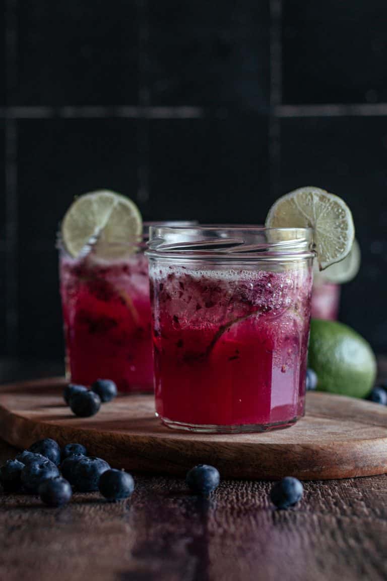 Two glasses of blueberry limeade on a wooden cutting board.