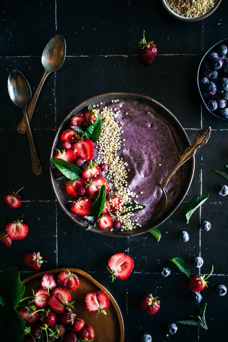 Blueberry smoothie topped with strawberries, pink blueberries, popped quinoa, on a black tile background surrounded by strawberries and blueberries.