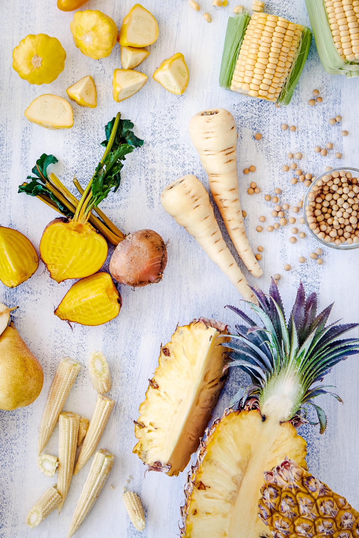 Distressed white surface covered in yellow foods including patty pan squash, sweet corn, beets, pineapple,  pear, and mini corn.
