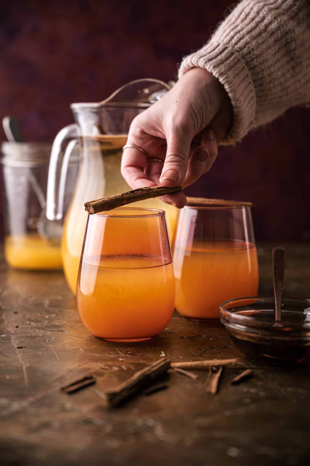 Two cups of pumpkin lemonade with a female hand placing a cinnamon stick on top of one glass.