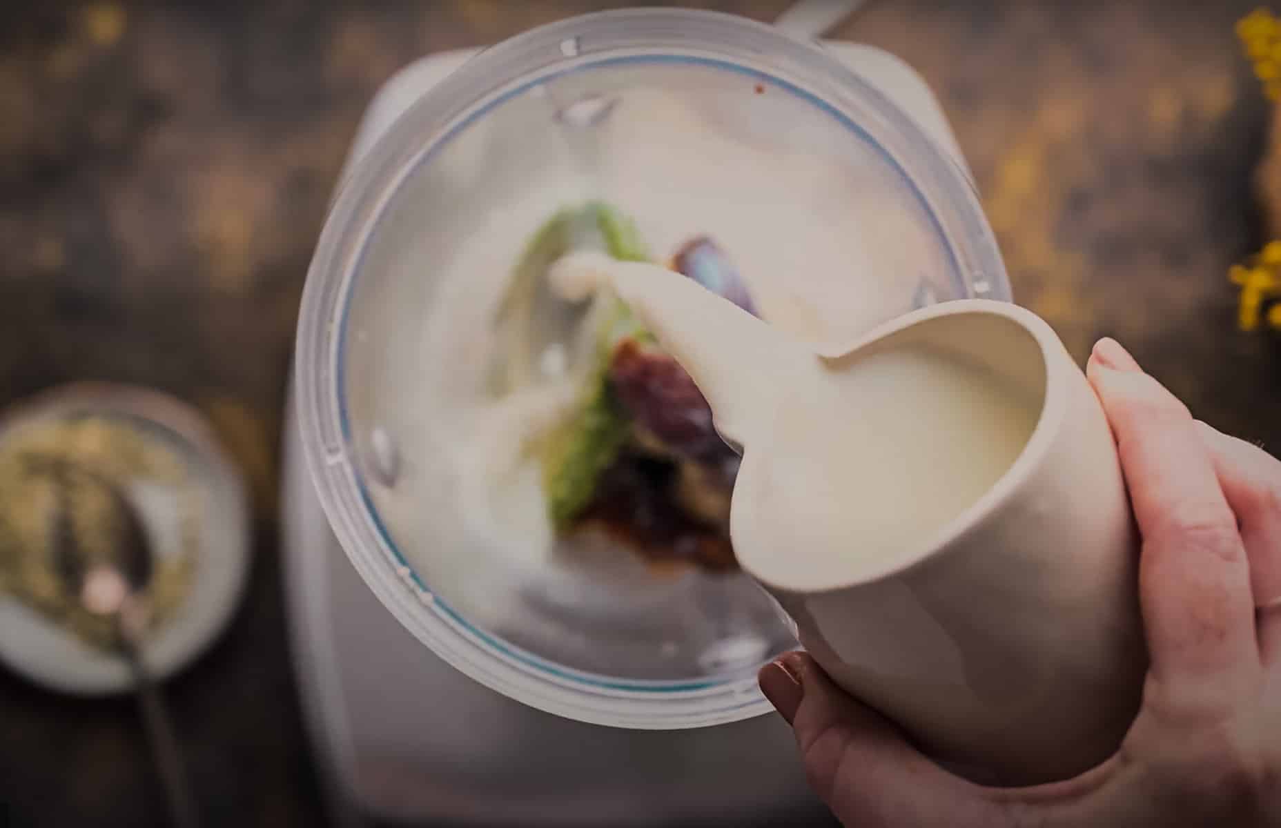 Hand pouring milk from a small jug into a blender.