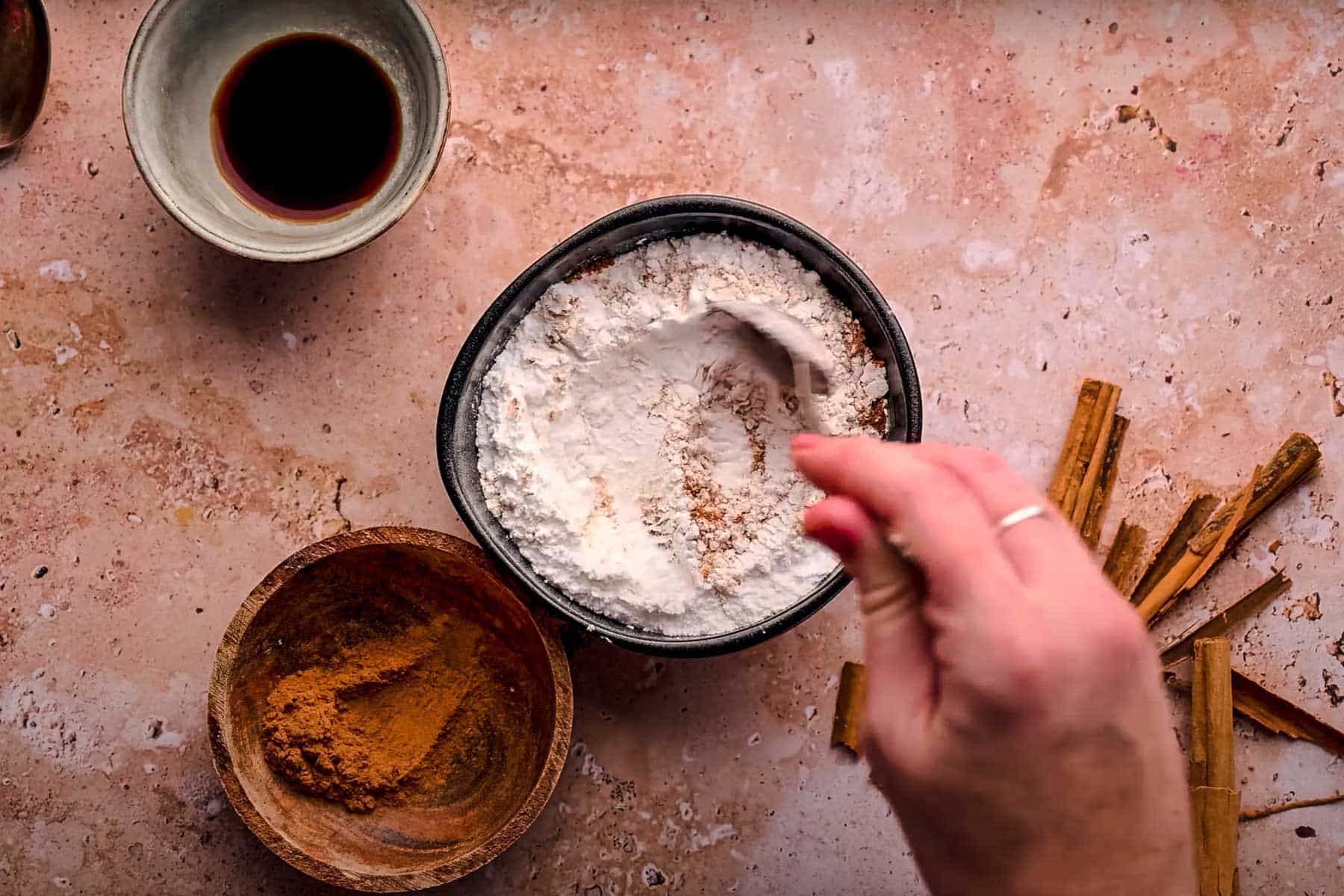 A hand stirs powdered sugar, cinnamon, and salt in a bowl with a spoon.