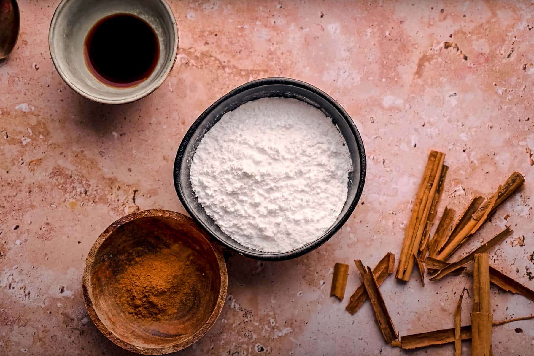 A flat lay image shows bowls of powdered sugar, cinnamon, and a vanilla extract, along with cinnamon sticks.