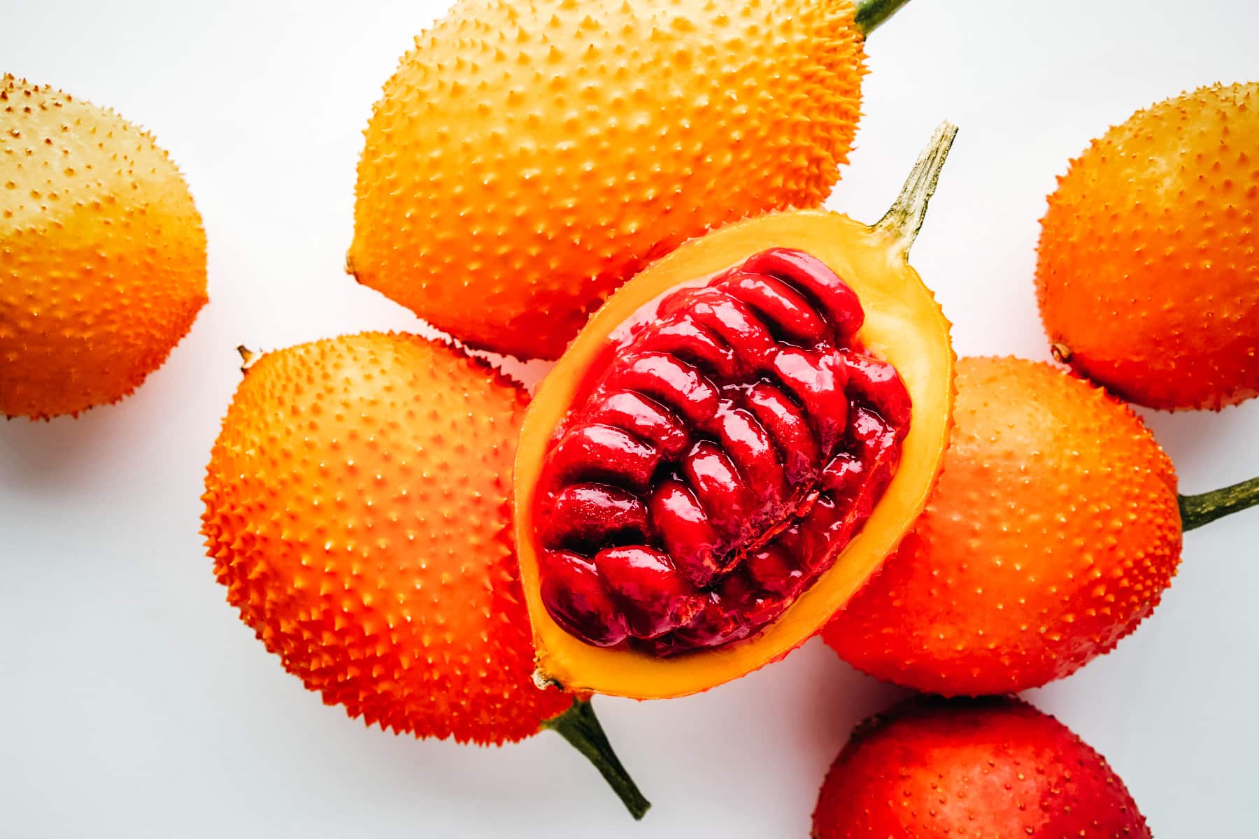 Several orange gac fruits with one cut in half showing its red colored seeds.