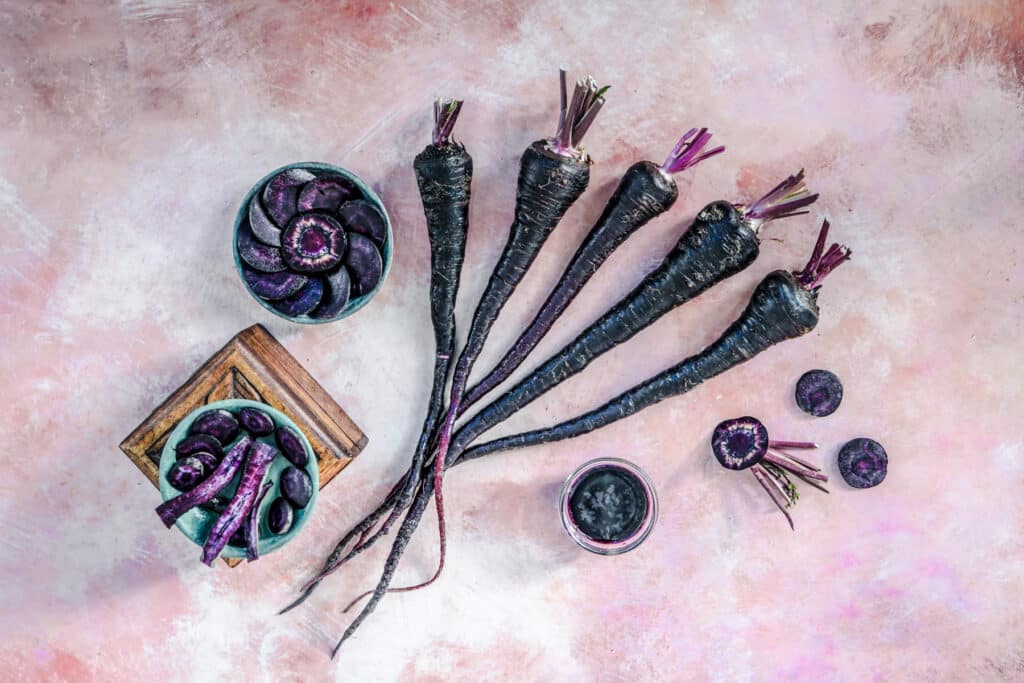 Full Pusa Asita black carrots along with cut slices on a white and pink surface.