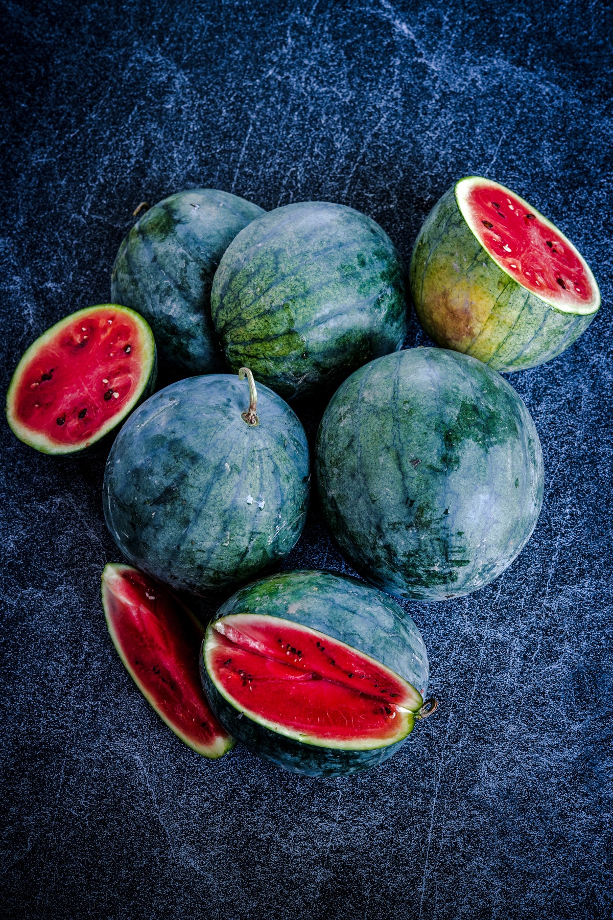 Blacktail mountain watermelon with dark green to. black skin and red flesh on a charcoal-navy blue surface.