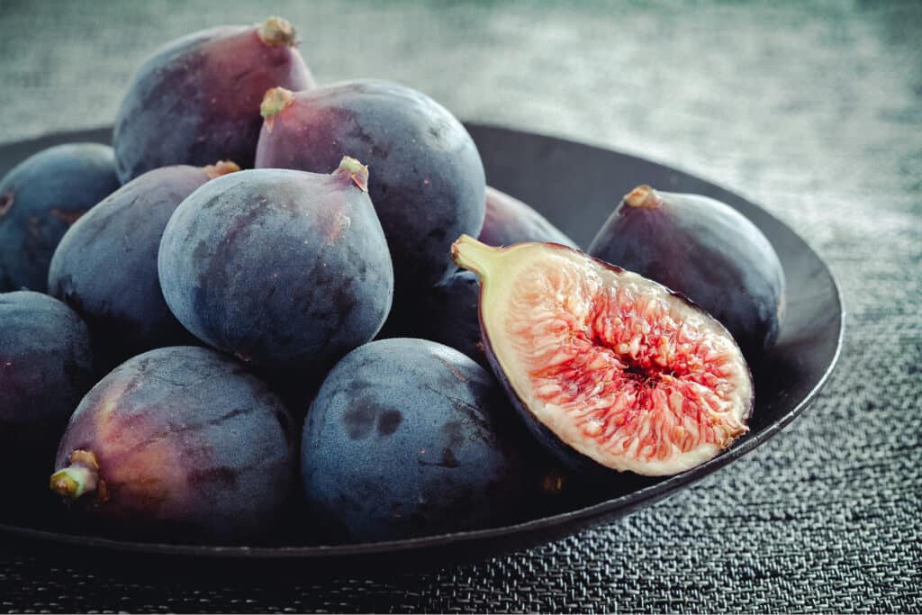 Black mission figs on a black plate with one cut in half.