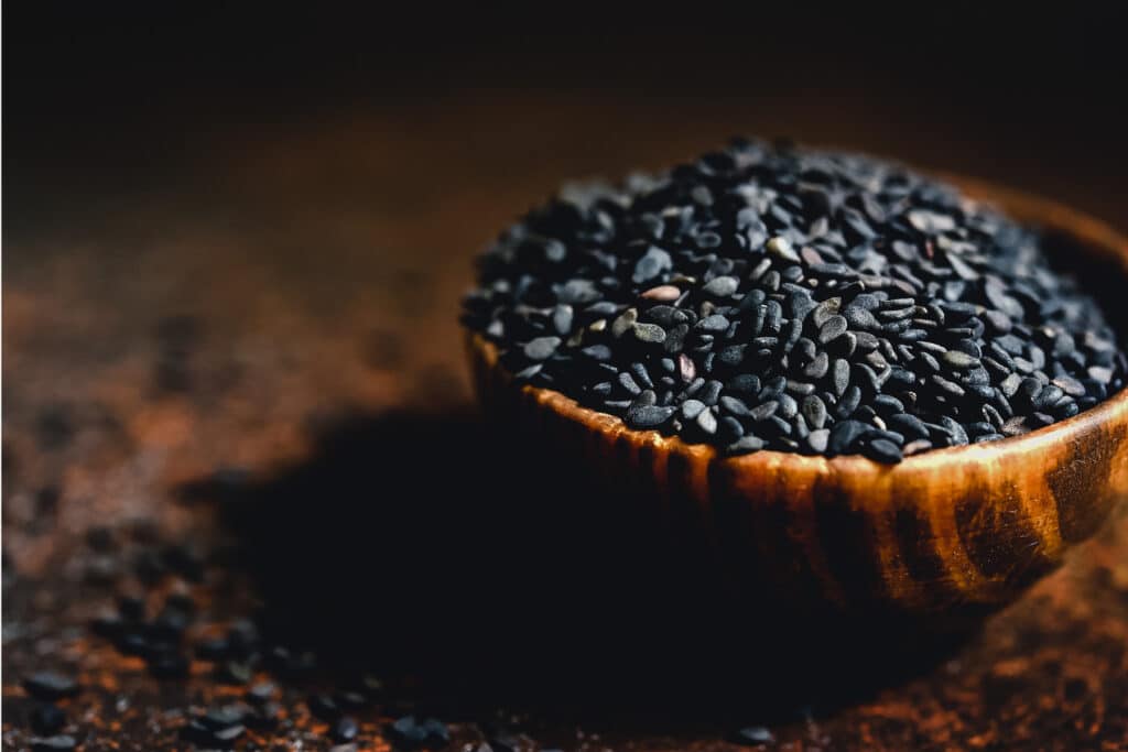 Black sesame seeds in a small wooden bowl overflowing onto the wooden table.