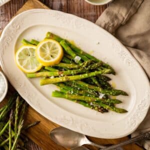 A white oval plate features perfectly cooked asparagus, delicately topped with lemon slices and a sprinkle of salt.
