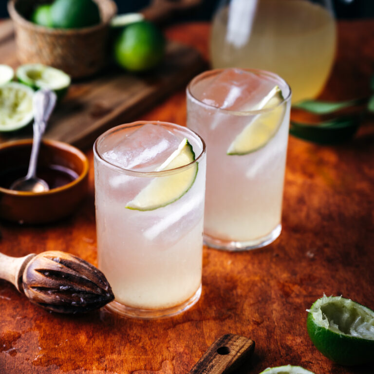 Two glasses of limeade with ice and lime wedges, on a wooden table surrounded by limes and a juicer.