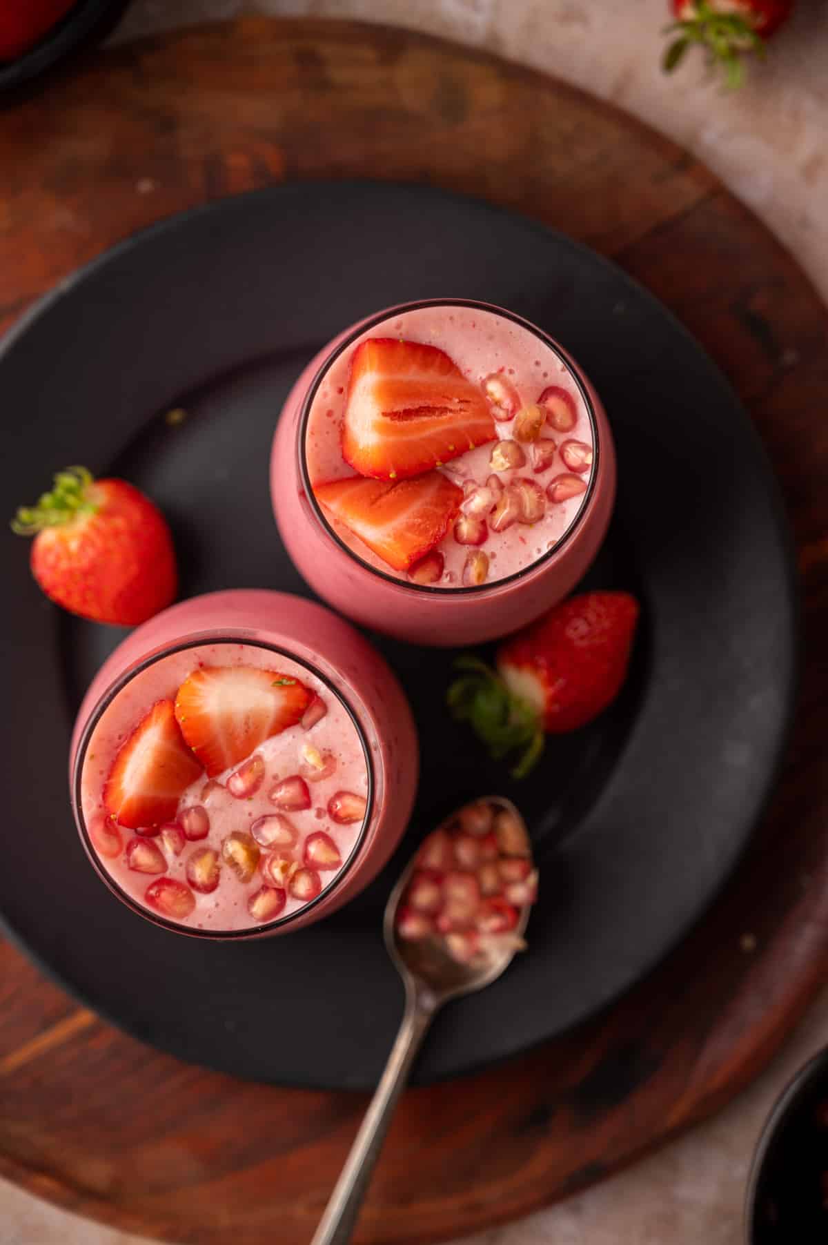 Two cups of pomegranate smoothie on a black plate with a spoon of arils next to it.