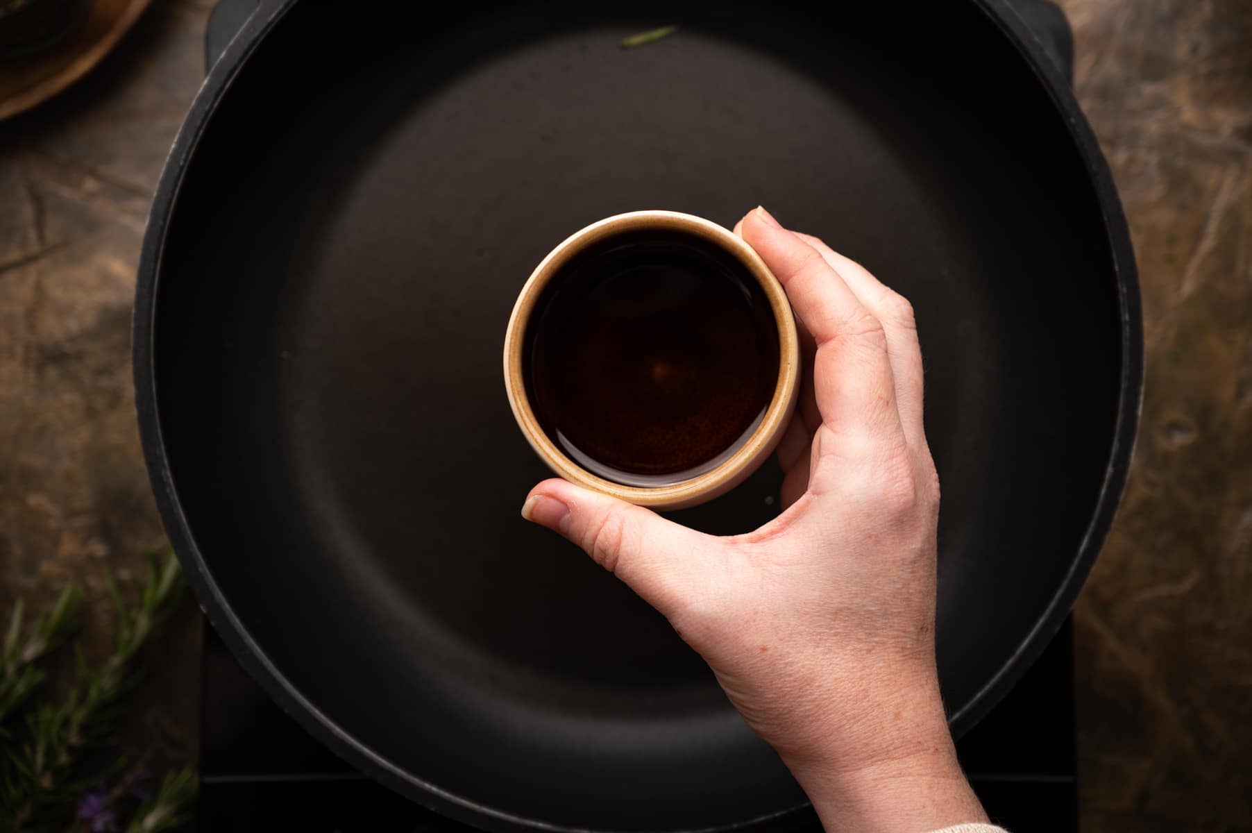 A hand holding a small cup of oil above an empty pan.