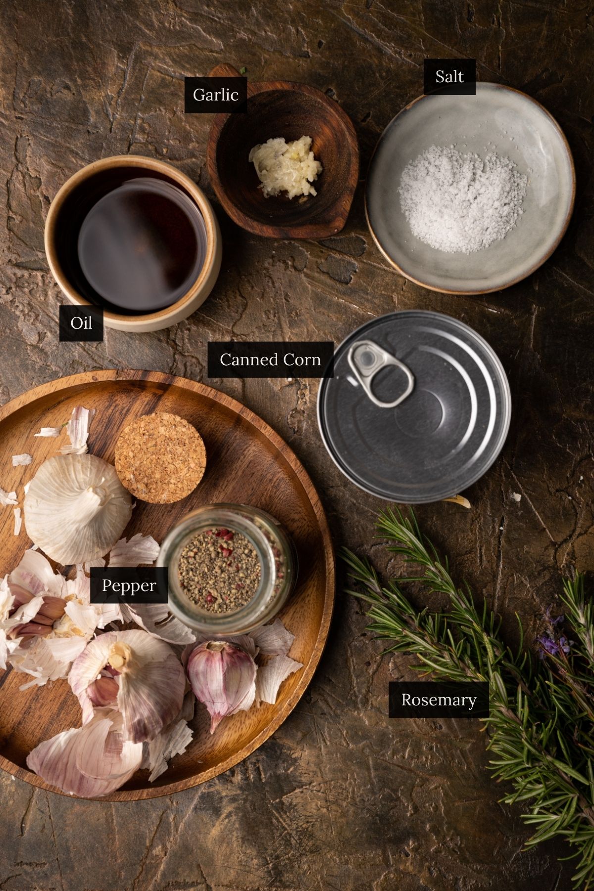 Overhead view of ingredients for canned corn on a textured surface: oil, canned corn, garlic, salt, garlic bulbs, pepper, and a rosemary sprig with labels.