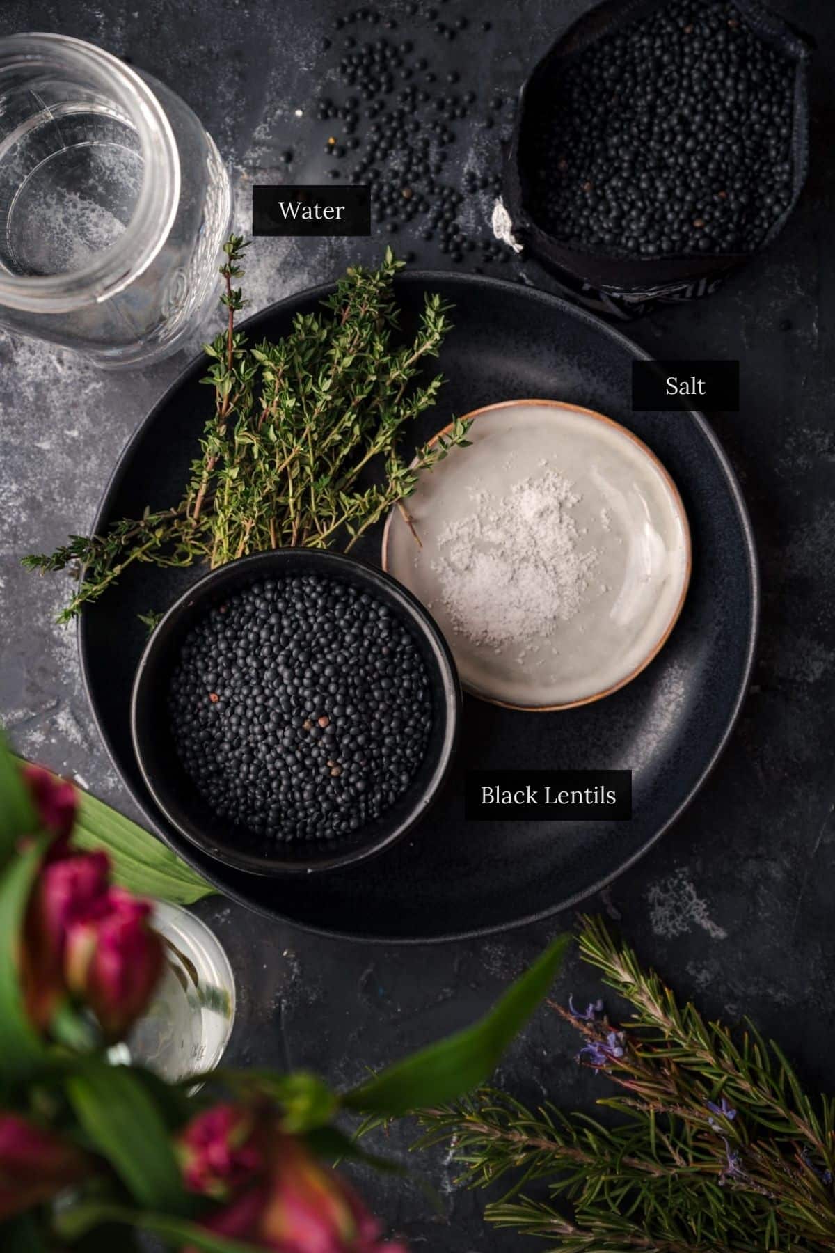 A dark plate with a bowl of black lentils, a bowl of salt, and sprigs of fresh herbs. A labeled jar of water and a bowl of black lentils are nearby.