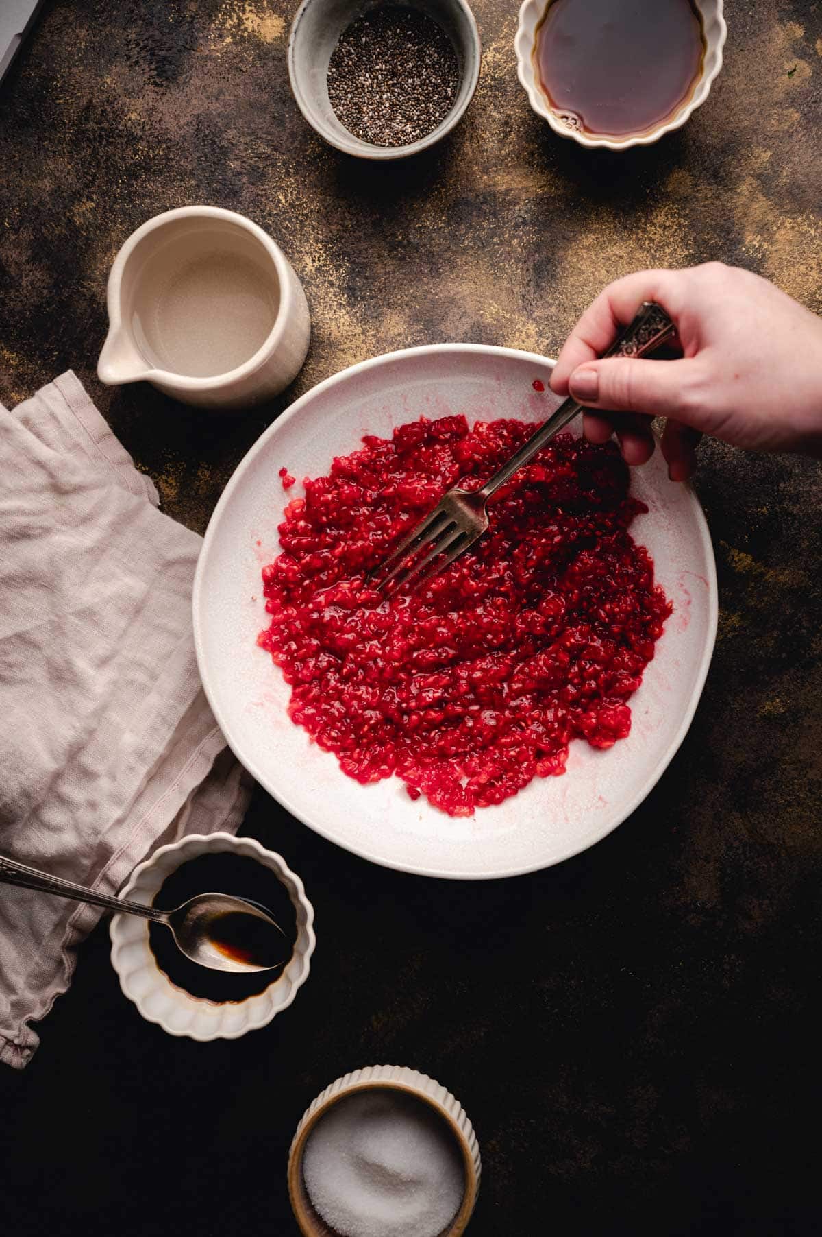 Fresh raspberries being smashed on a white plate with ingredients in bowls surrounding it.