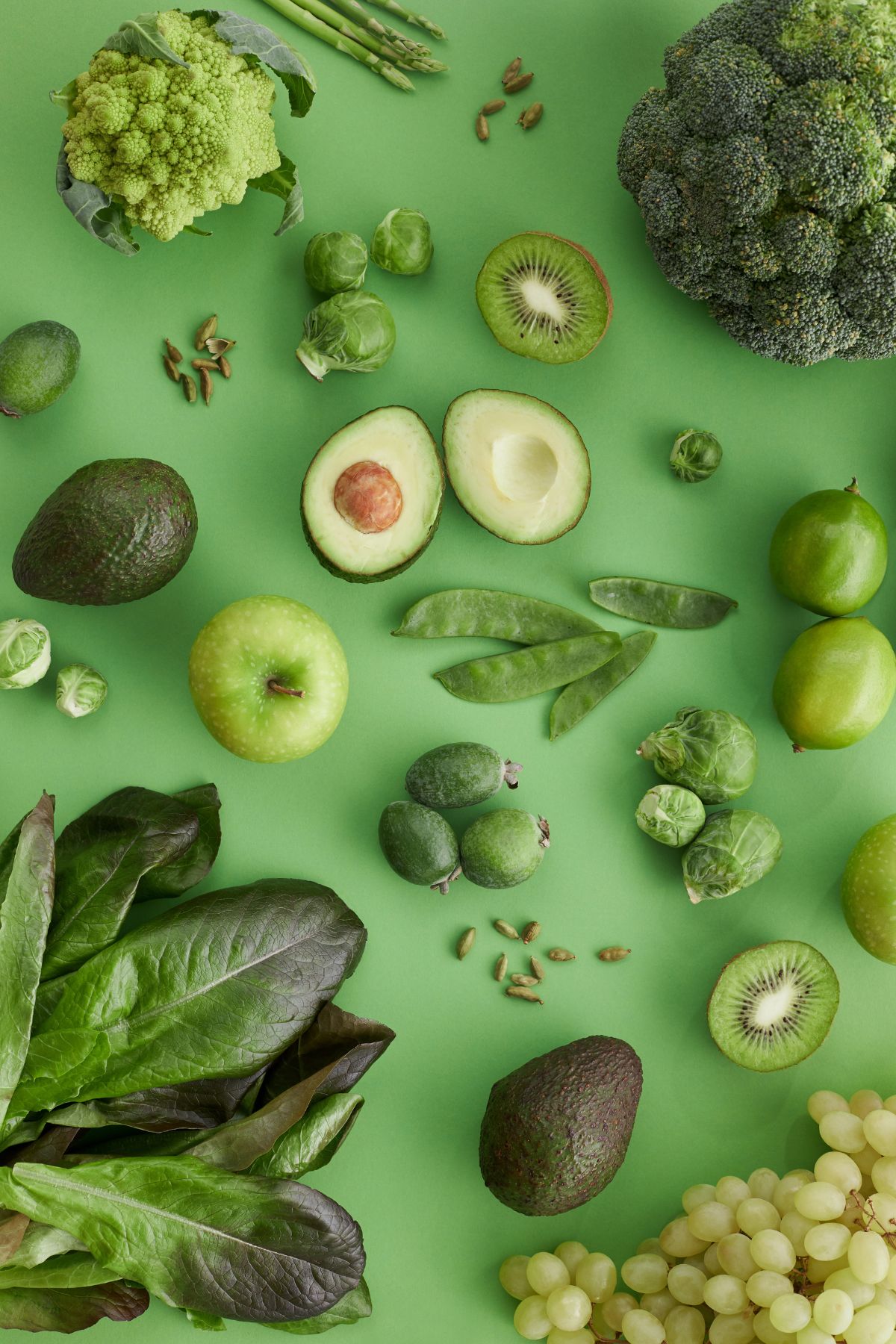 A variety of fruits that are green, like avocado, kiwi, apple, and green vegetables such as broccoli, Brussels sprouts, grapes, and lettuce are artfully arranged on a vivid green background.