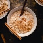 A bowl of oatmeal topped with milk, a sprinkle of brown sugar cinnamon, with a wooden spoon and cinnamon sticks placed around the bowl.