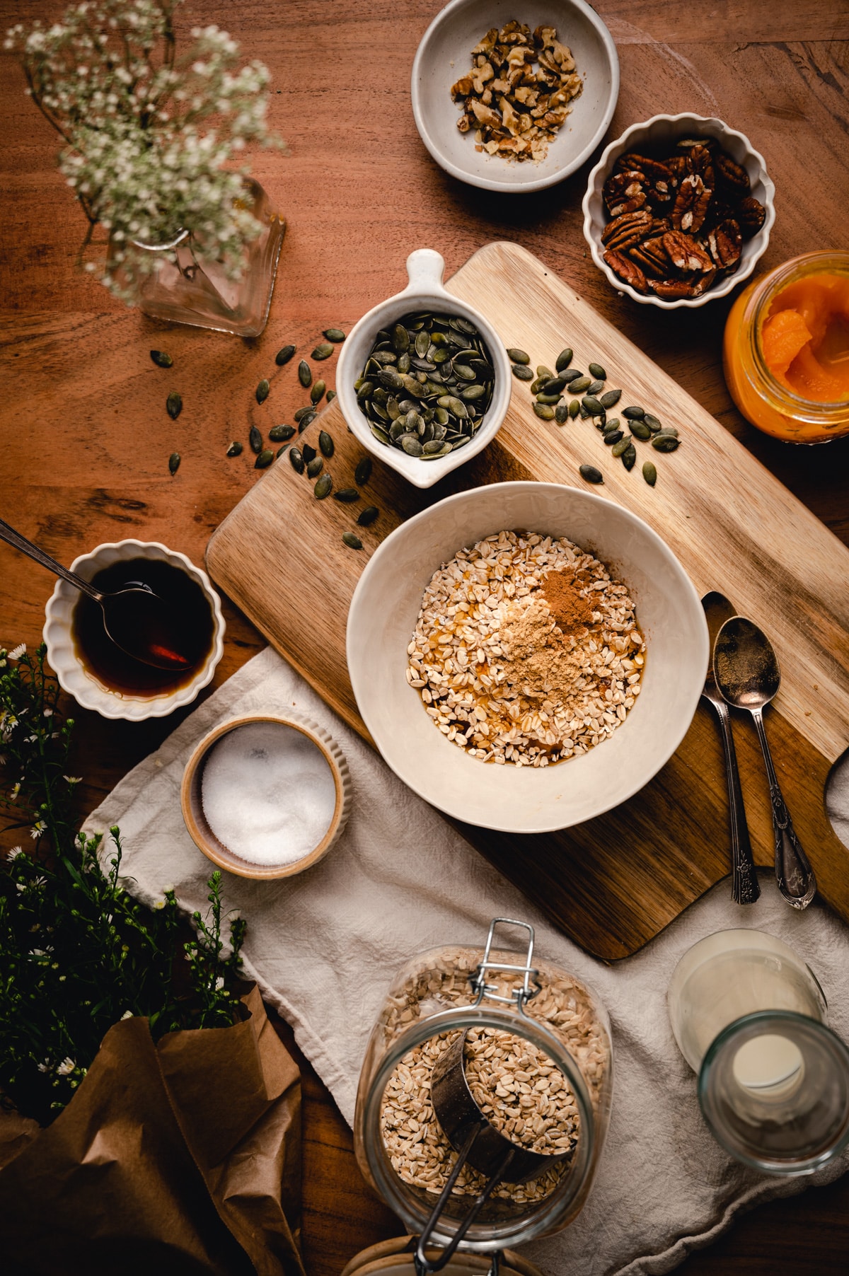 A wooden table with a bowl of oats, jar of oats, pumpkin seeds, nuts, a jar of maple syrup, and a small dish of syrup.