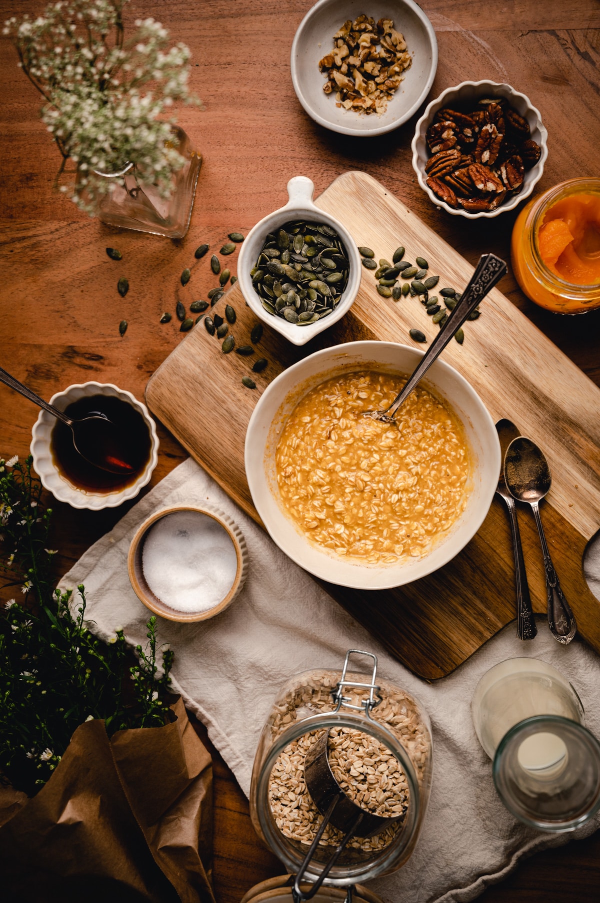 A bowl of oatmeal on a wooden board, surrounded by seeds, nuts, a jar of oats, and a bottle of milk, on a rustic table.