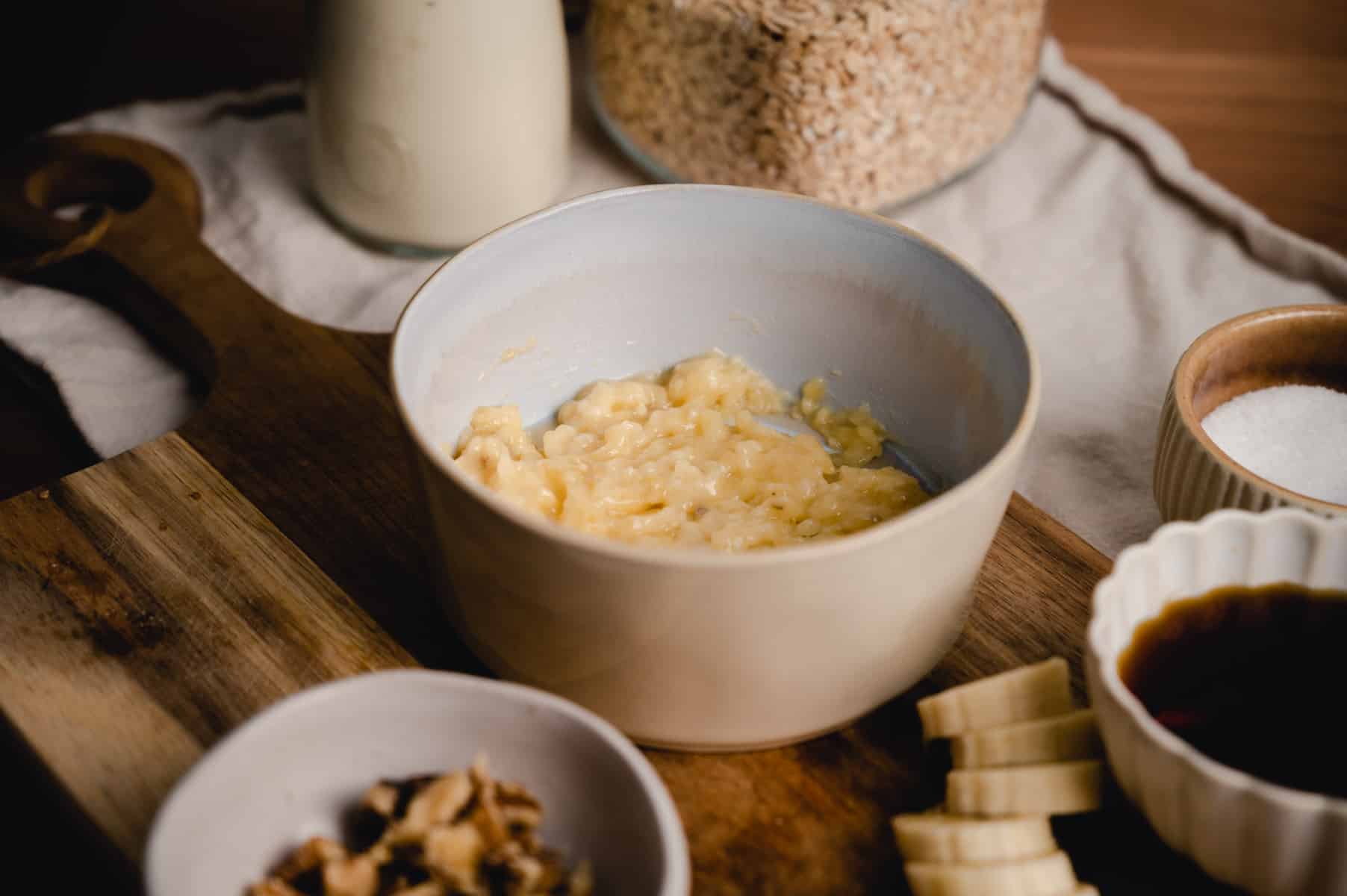A bowl containing a mashed banana mixture is placed on a wooden cutting board.