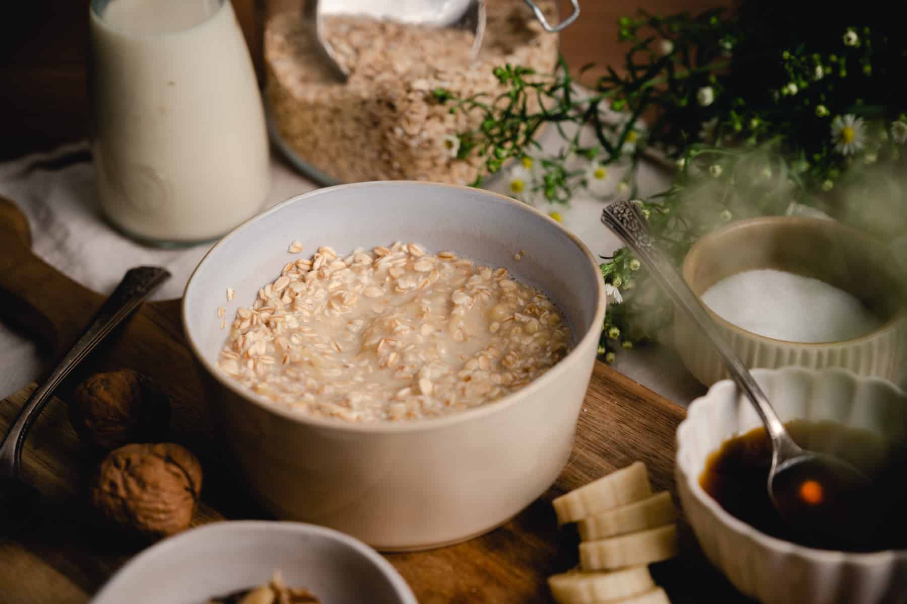 A bowl of oatmeal surrounded by ingredients including milk, rolled oats, walnuts, banana slices, and a small bowl of a maple syrup.