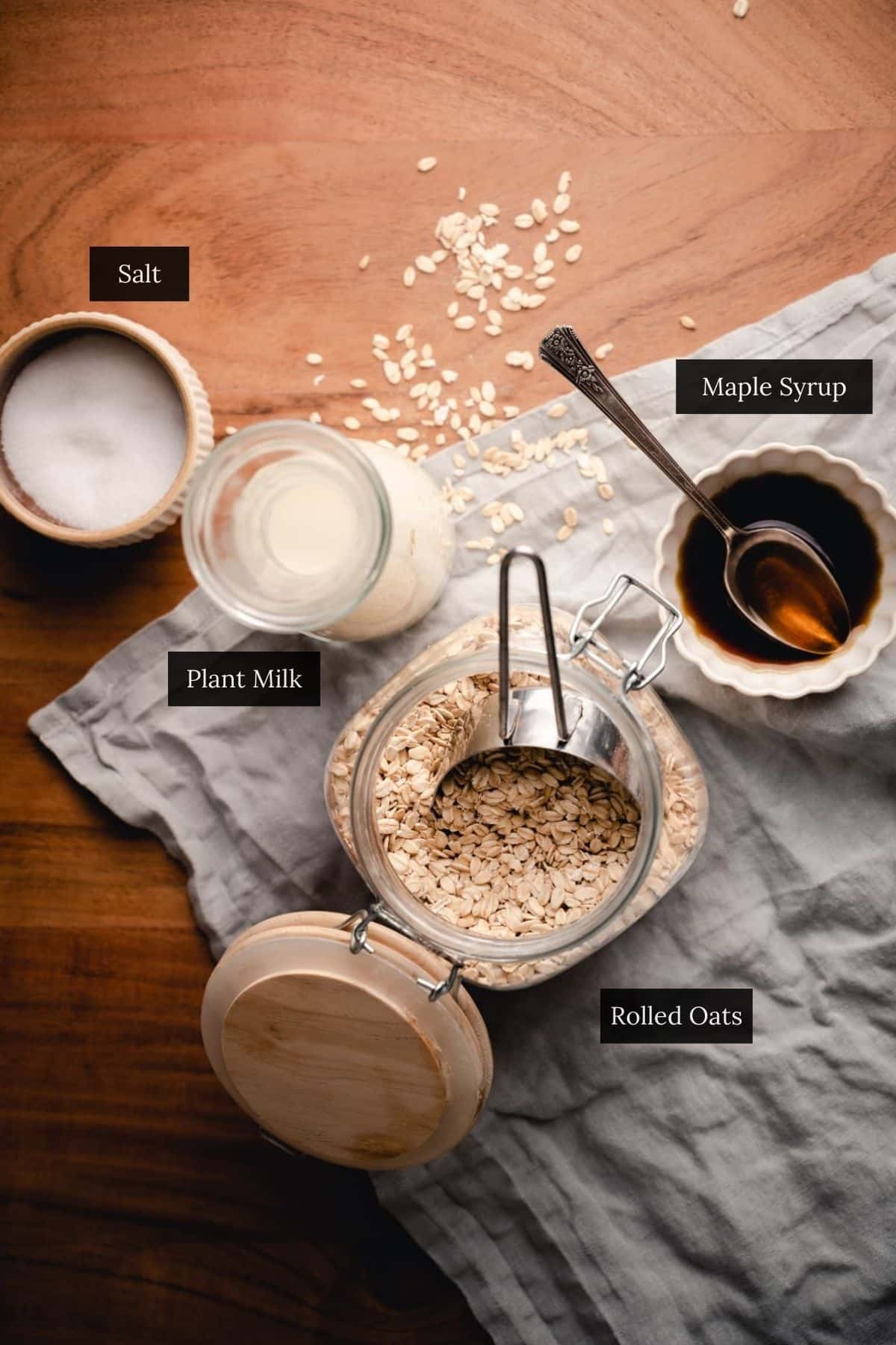 A jar of rolled oats with a scoop, a bowl of maple syrup with a spoon, a small jar of plant milk, and a container of salt sit on a wooden surface with a gray cloth.