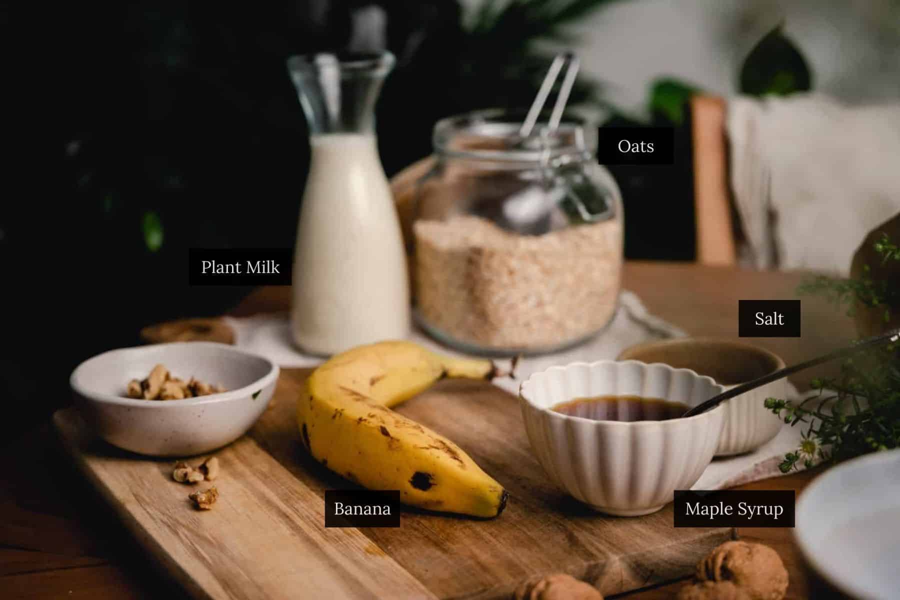 A breakfast setup featuring a banana, plant milk, a bowl of maple syrup, a bowl of nuts, a jar of oats, and a bowl of salt on a wooden board.