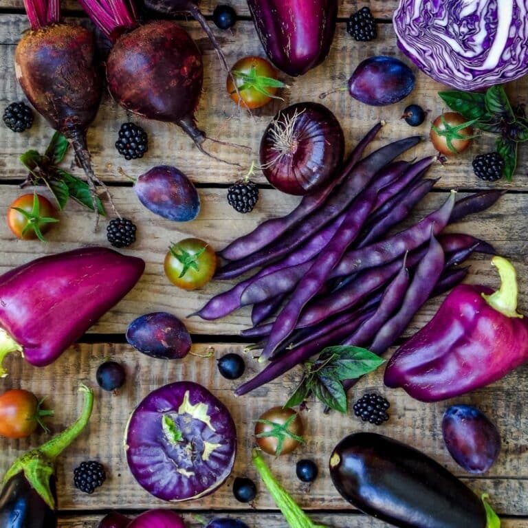 A vibrant selection of foods that are purple, including eggplant, cabbage, peppers, beans, tomatoes, plums, beets, and blackberries, artfully arranged on a wooden surface.