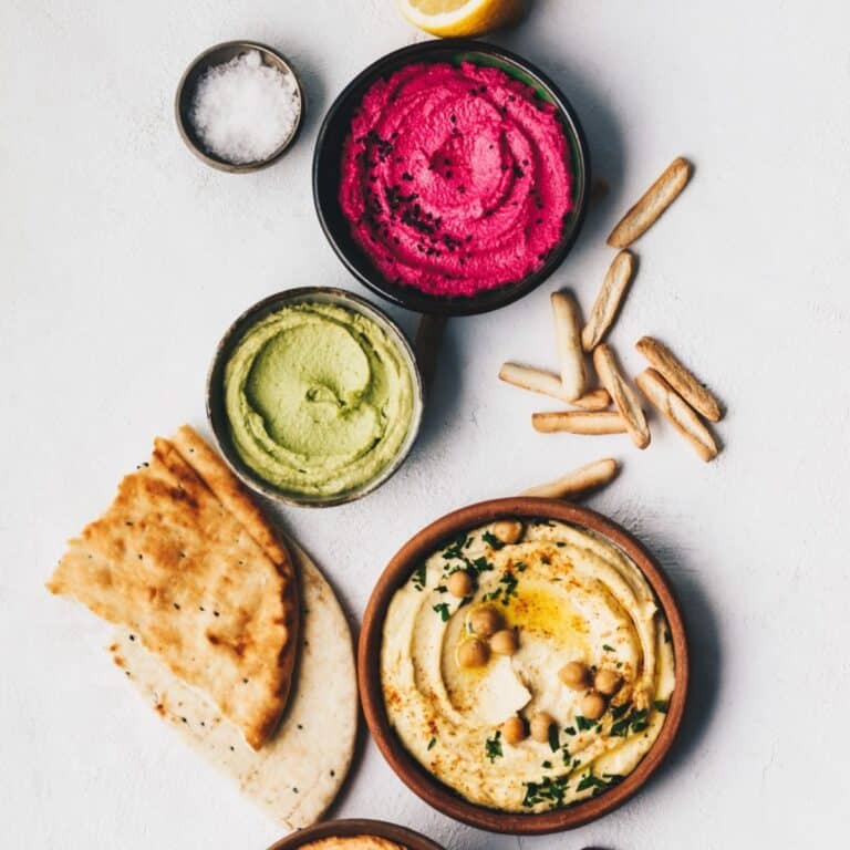 Assorted hummus dishes in bowls on a white surface, served with hummus along with crunchy breadsticks, soft pita bread, and a lemon wedge.