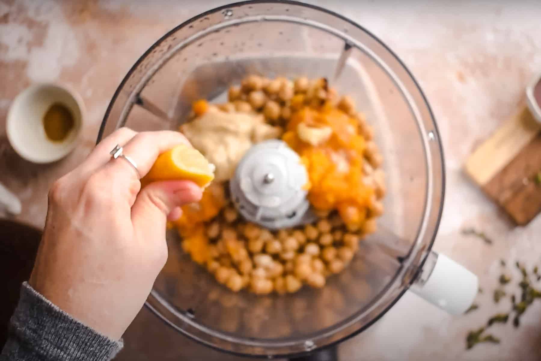 A hand squeezes a lemon slice into a food processor containing chickpeas, tahini, oil, and pumpkin.