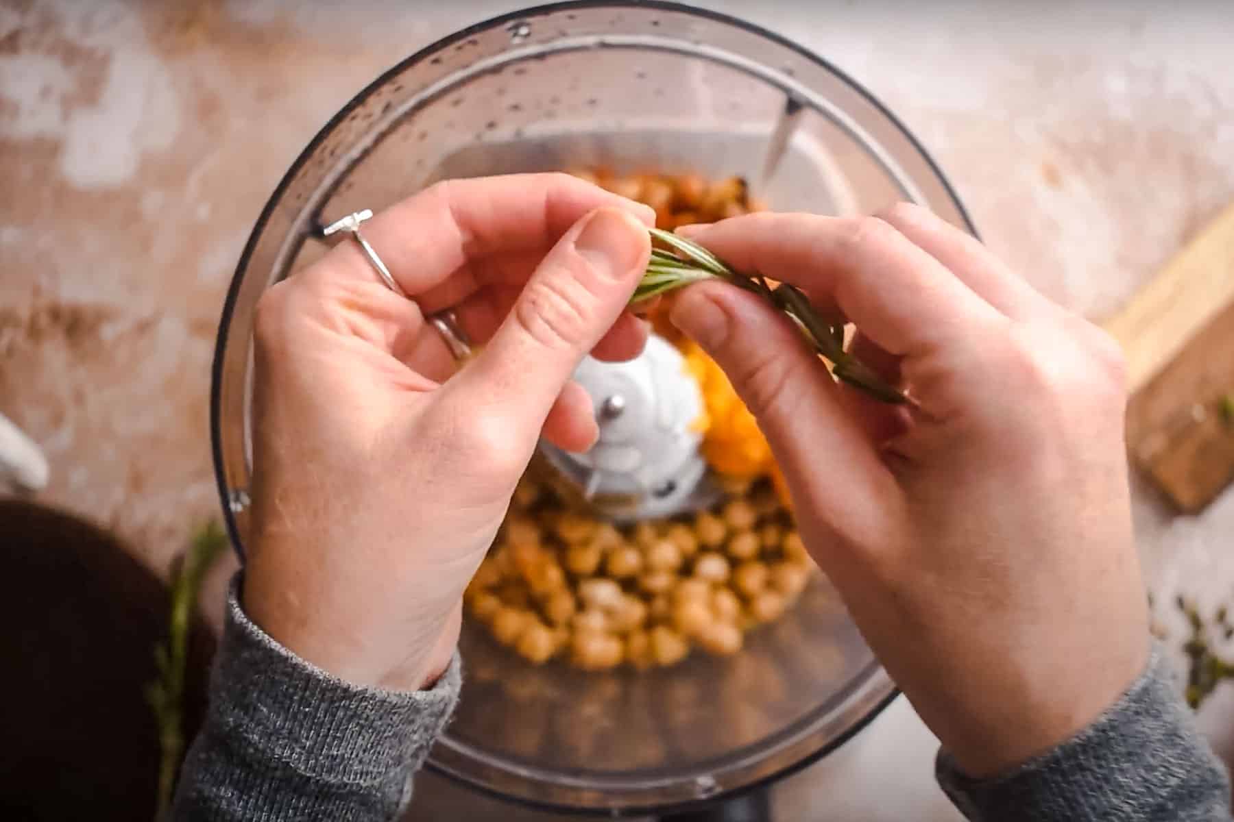 Hands adding fresh herbs to a food processor containing chickpeas, crafting a delightful pumpkin hummus.