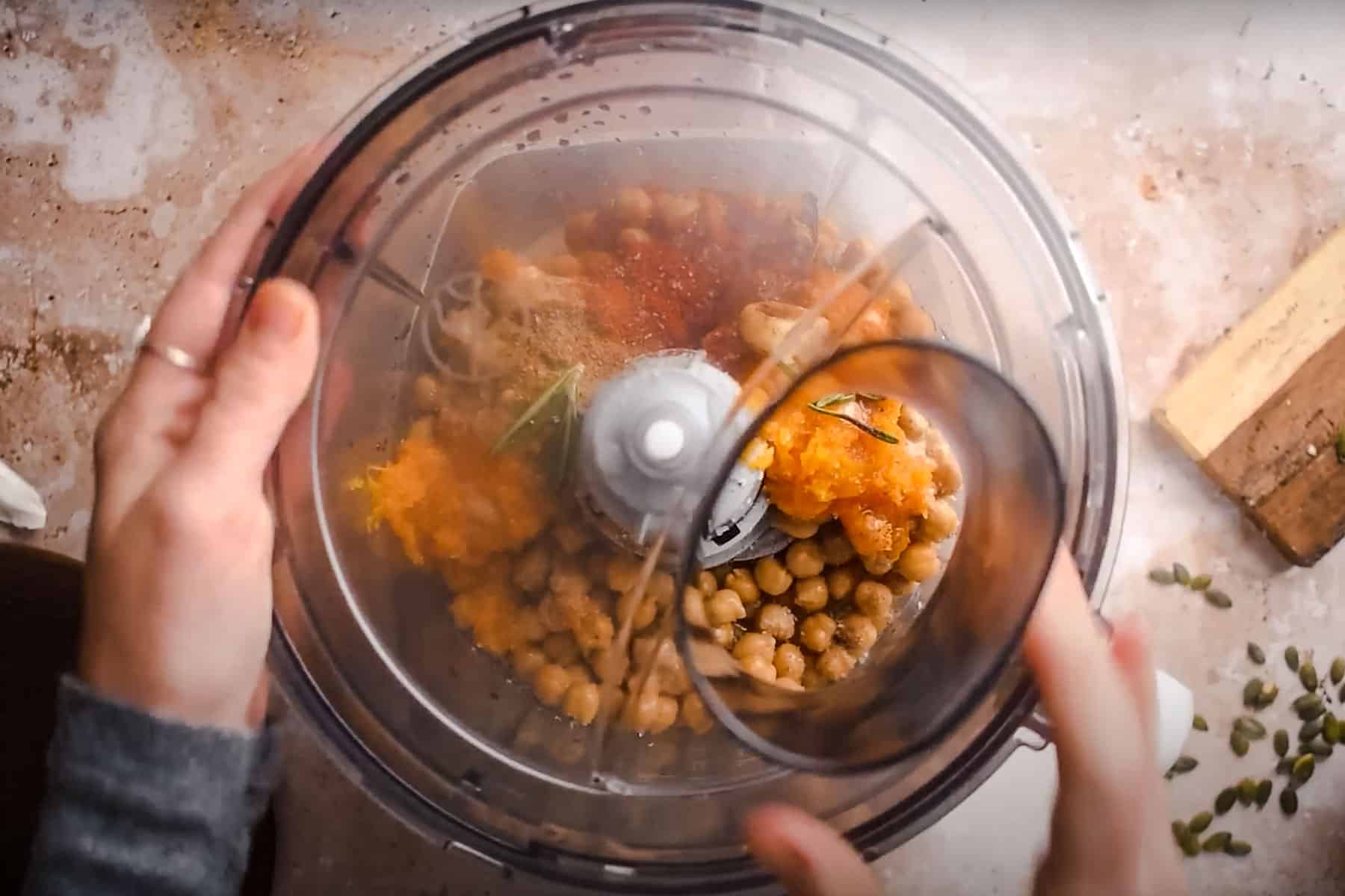 Top view of a person placing a lid on a food processor containing chickpeas, pumpkin chunks, spices, and a rosemary sprig.