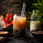 A glass of bubble tea with tapioca pearls and ice, served with a clear straw, surrounded by tea ingredients, sugar, a jar of milk, and a red flower in the background—nothing beats this classic milk tea recipe.