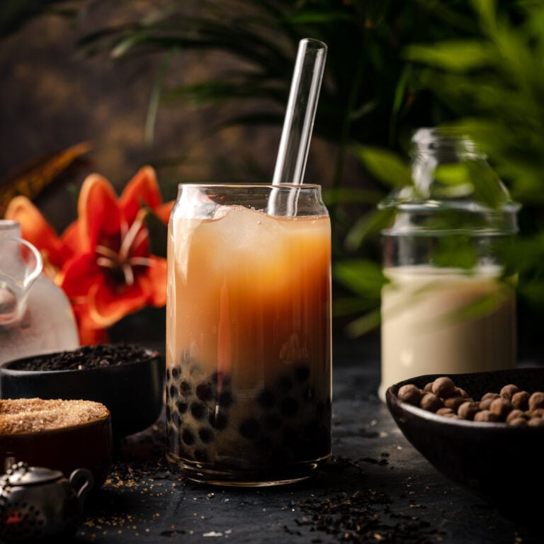 A glass of bubble tea with tapioca pearls and ice, served with a clear straw, surrounded by tea ingredients, sugar, a jar of milk, and a red flower in the background—nothing beats this classic milk tea recipe.