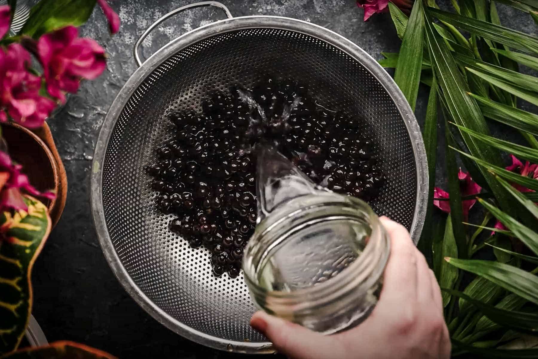 A hand pours water from a jar into a metal colander filled with black tapioca pearls.
