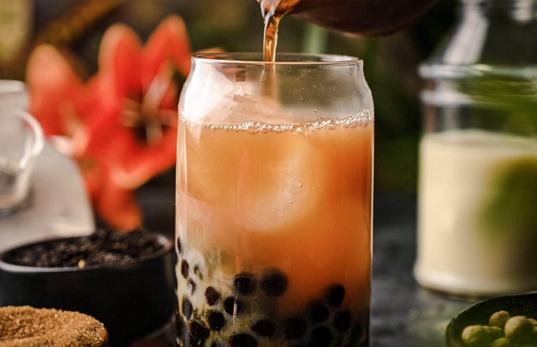 A glass of bubble tea is being filled with a brown liquid from a container, showcasing a classic milk tea recipe. The drink contains tapioca pearls. In the background, there's a small bowl of loose leaf tea, a jar, and a flower.