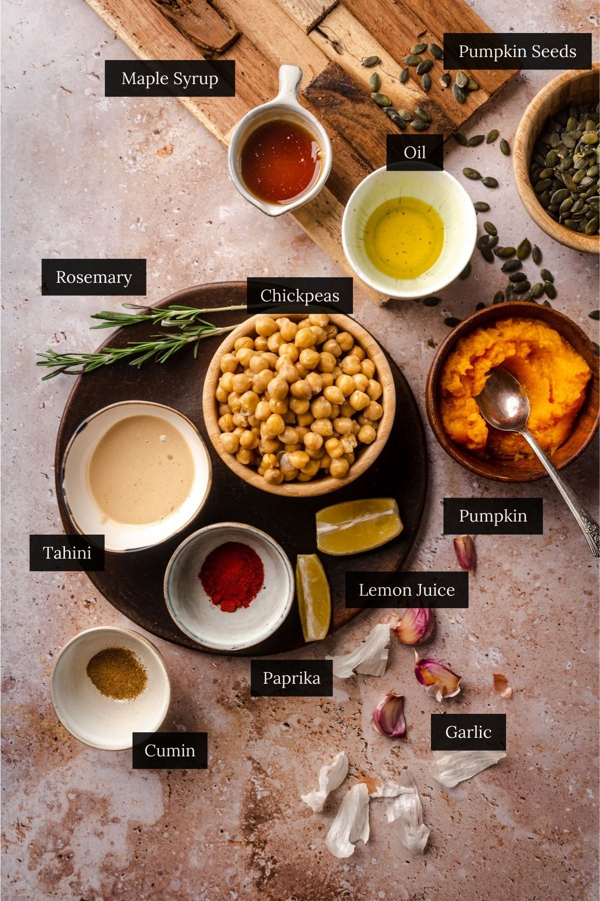 Top-down view of various ingredients labeled, including chickpeas, pumpkin, pumpkin seeds, oil, maple syrup, rosemary, tahini, lemon juice, paprika, cumin, and garlic arranged on a textured surface.