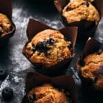 Several freshly baked vegan blueberry muffins in brown paper liners on a dark surface with scattered blueberries.
