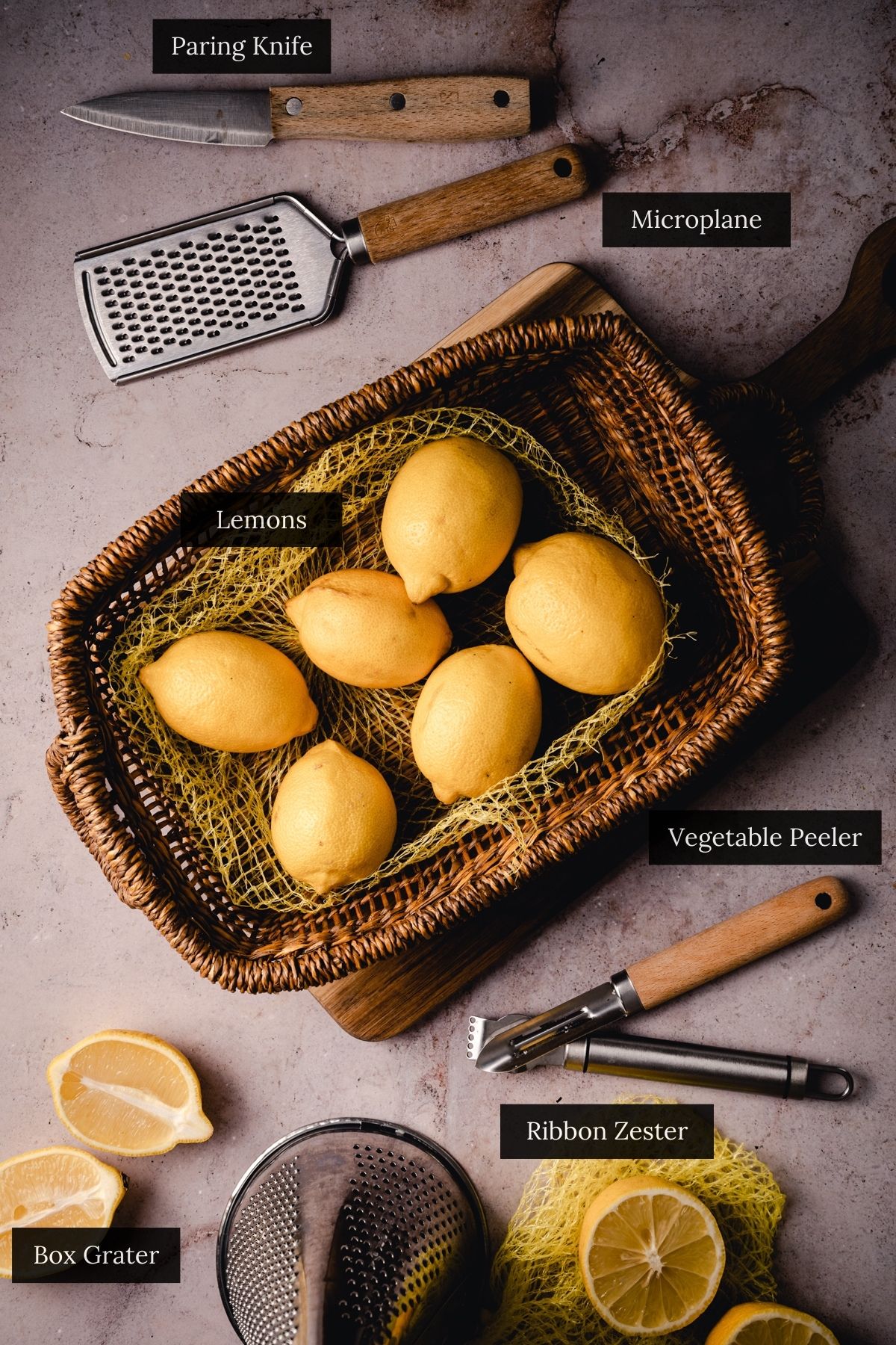 Wicker basket with six lemons sits next to a box grater, microplane, paring knife, vegetable peeler, and ribbon zester.