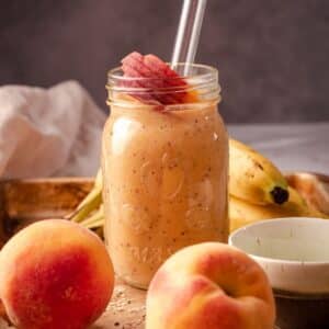 Peach smoothie in a mason jar on a tray with bananas and peaches.