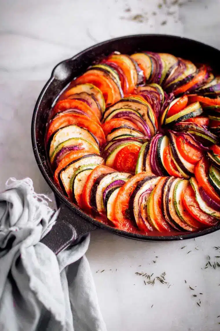 A round black baking dish filled with neatly arranged slices of zucchini, eggplant, and tomato, garnished with herbs; a beautiful vegan casserole. A grey cloth is placed next to the dish on a light-colored surface.