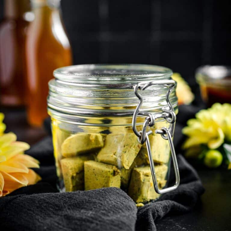 A glass jar filled with vegetable bouillon sits on a black cloth, surrounded by yellow flowers and bottles of veggie broth.