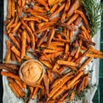A tray of roasted sweet potato fries garnished with rosemary and thyme, accompanied by a small dish of dipping sauce.