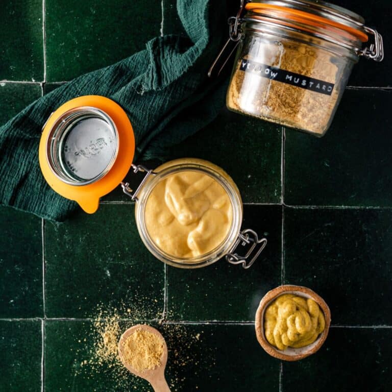 Jars of mustard on a dark green tile surface next to a wooden spoon with mustard powder and a small bowl of dijon mustard.