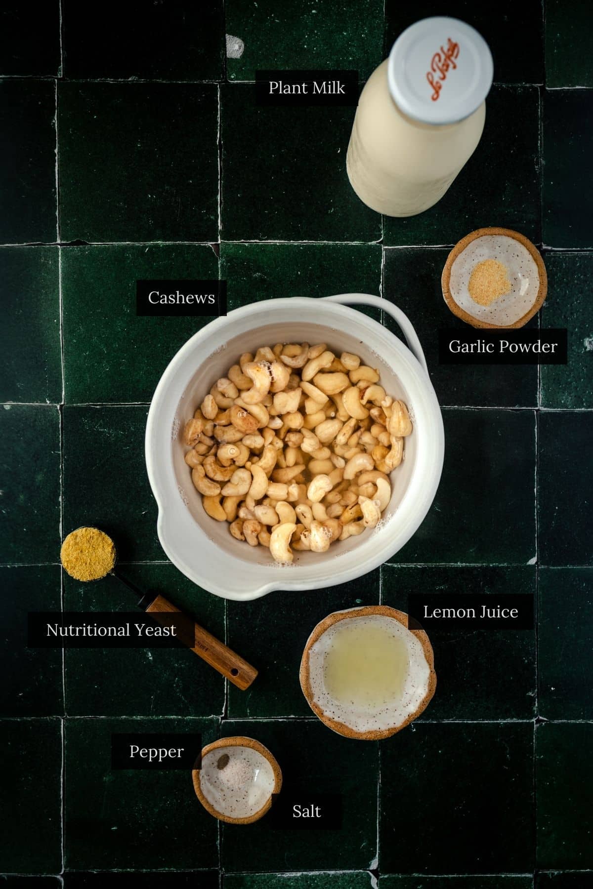 Top view of ingredients on a dark tiled surface: plant milk, cashews, garlic powder, lemon juice, salt, pepper, and nutritional yeast.