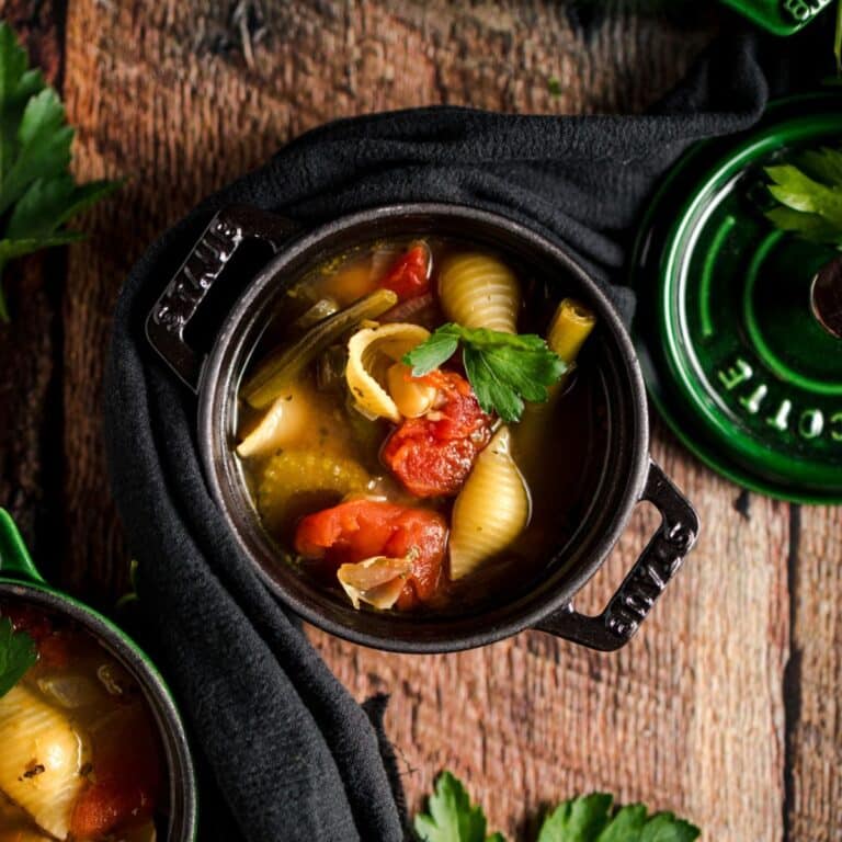 Small black pot with minestrone soup featuring pasta shells, tomatoes, and herbs, garnished with parsley.