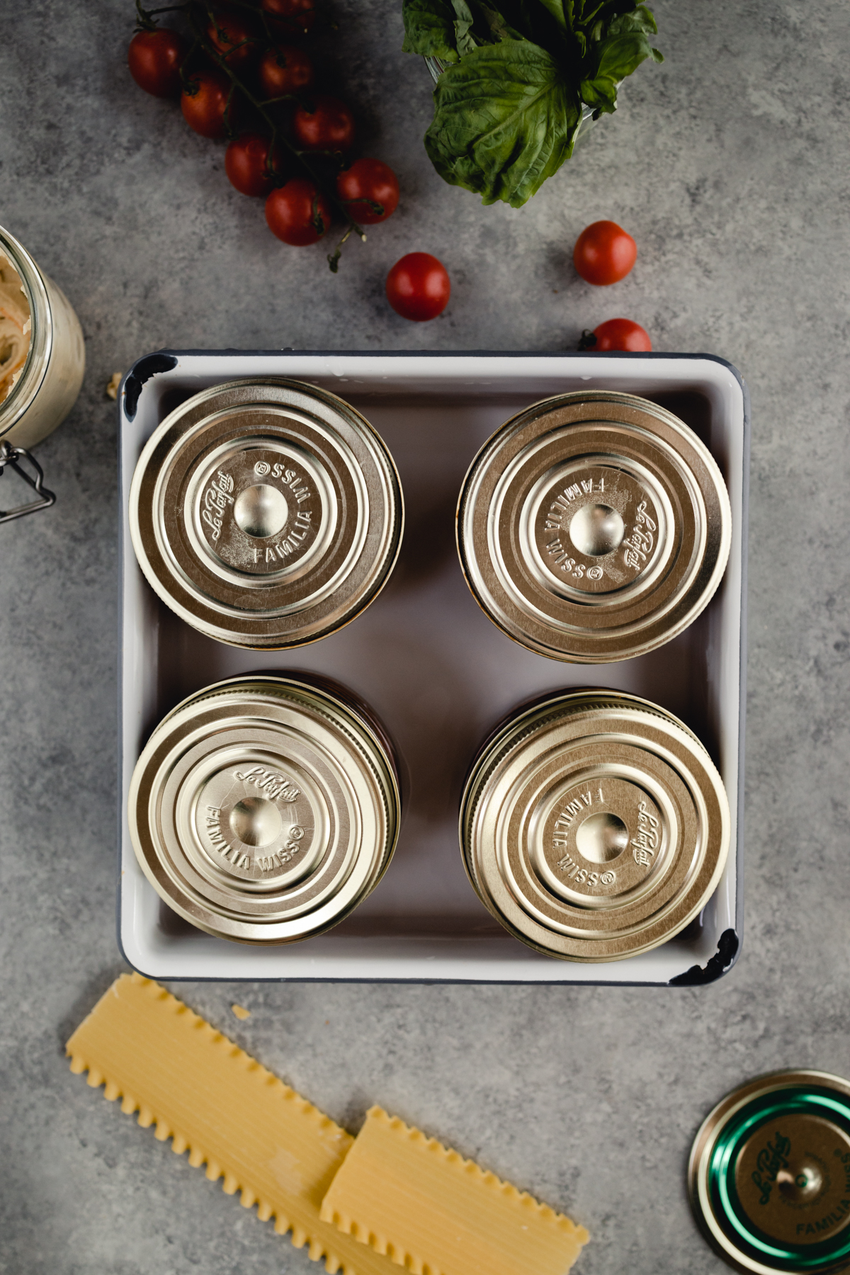 Prepared individual sized lasagnas in glass jars in a 9 by 9 pan with water with lids rested on top.